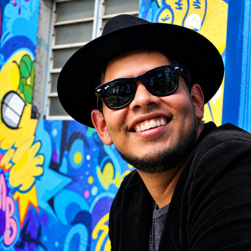 Victor Renan, de chapéu e óculos escuros, sorrindo em frente a um muro grafitado
